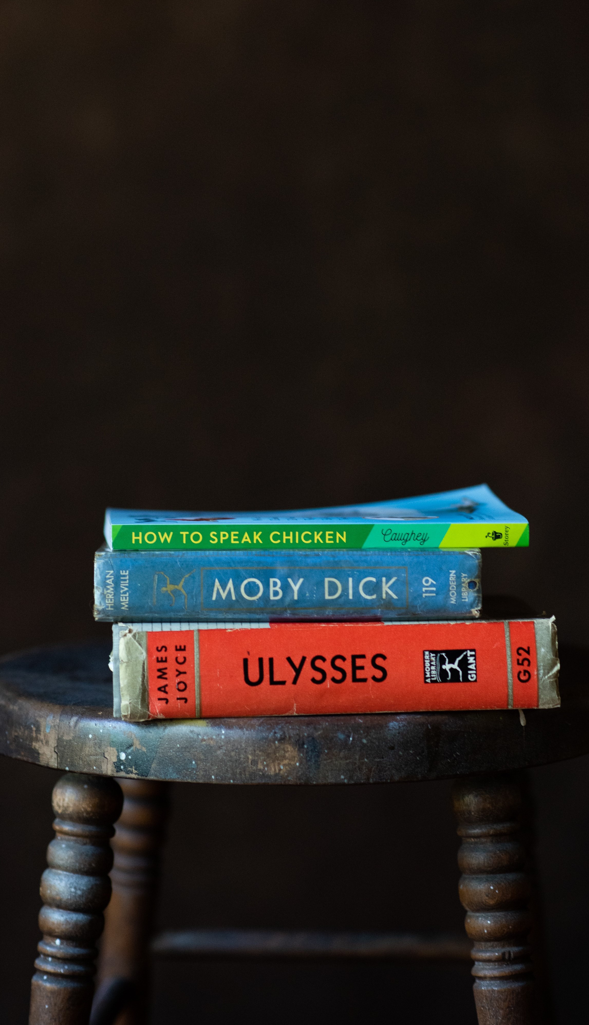 A stack of three books on a stool. The books are How To Speak Chicken by Caughey, Moby Dick by Herman Melville, and Ulysses by James Joyce.