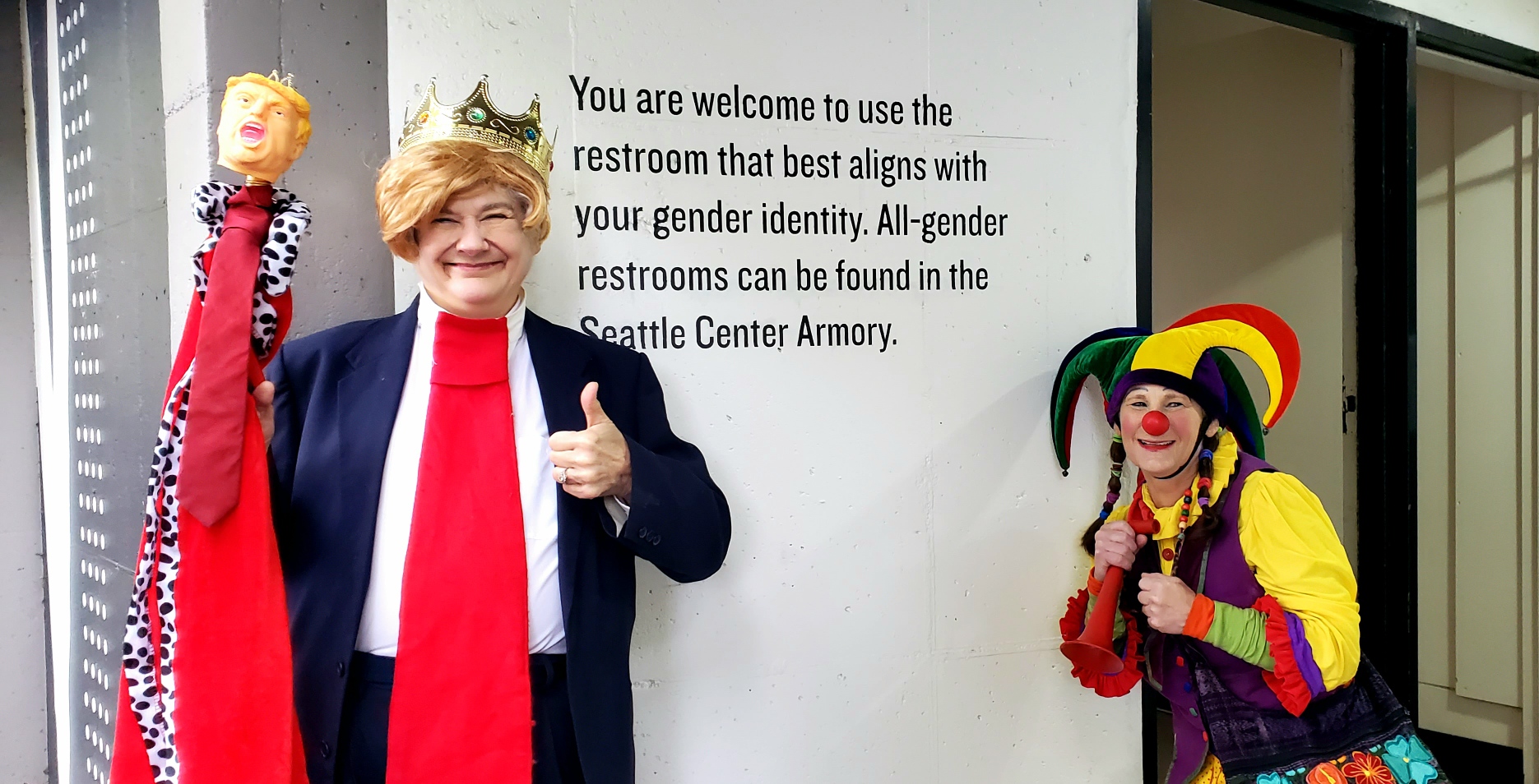 Victoria Millard as a Trump clown thumbs up next to all-gender restrooms sign