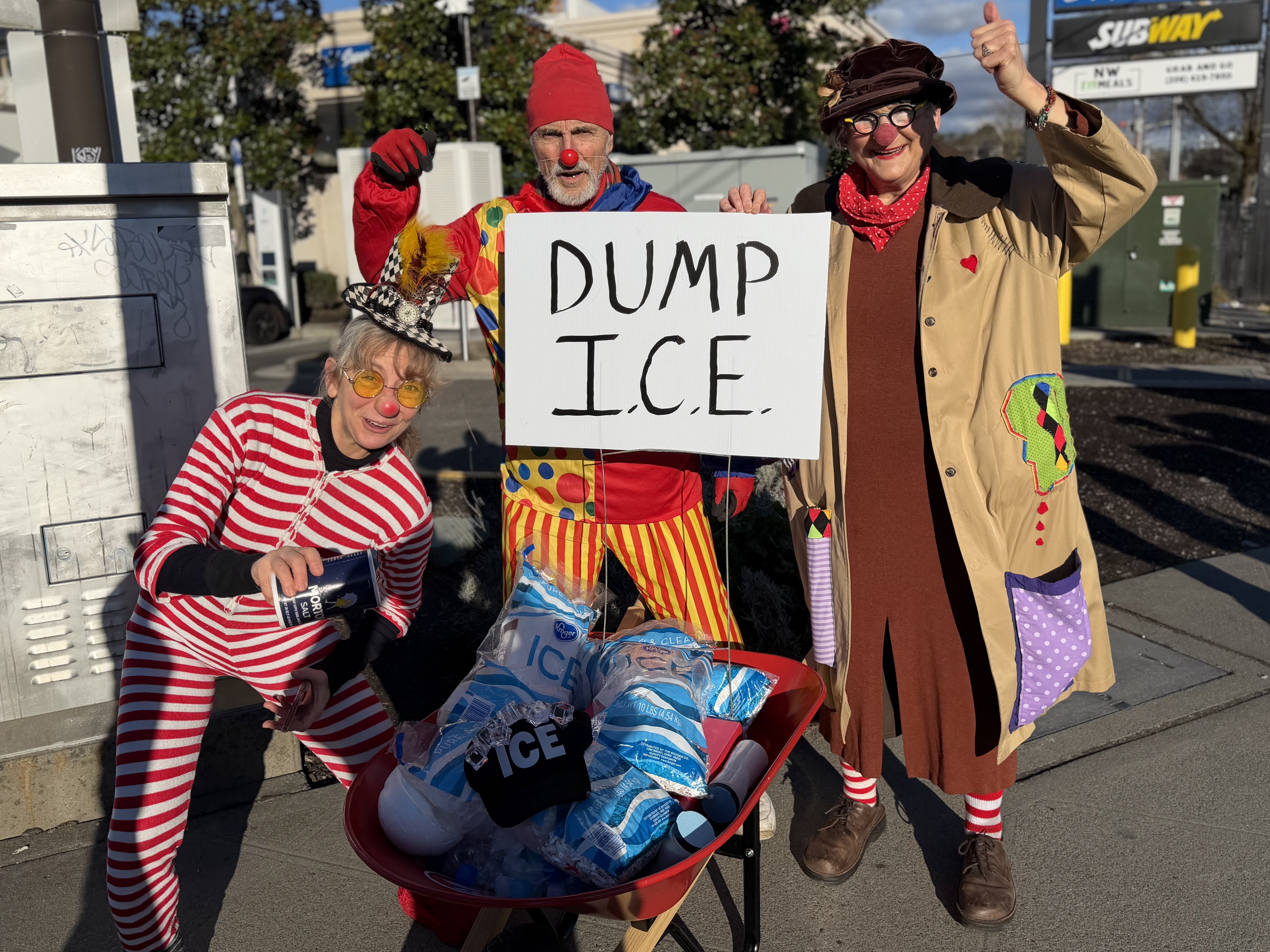 Clowns push a wheelbarrow full of ice with a sign that reads "Dump I.C.E."