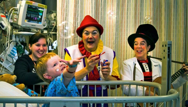 Victoria Millard as Dr. La Fou blows bubbles as another clown plays a guitar, a child in a hospital crib reaches for a bubble, and the child's mom looks on smiling.