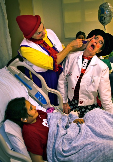 Victoria Millard as Dr. La Fou pulls a scarf from another clown's ear as a child in a hospital bed smiles