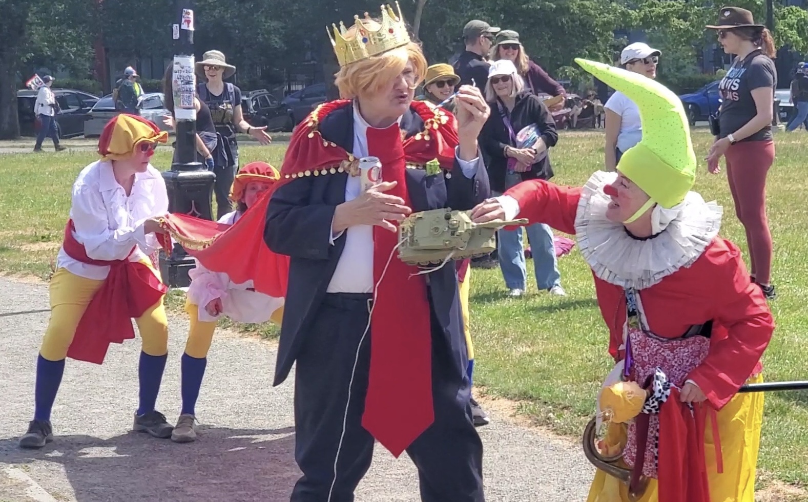 Victoria Millard at No Kings protest dressed as clown Trump with a crown, holds toy tank while other clowns hold her train