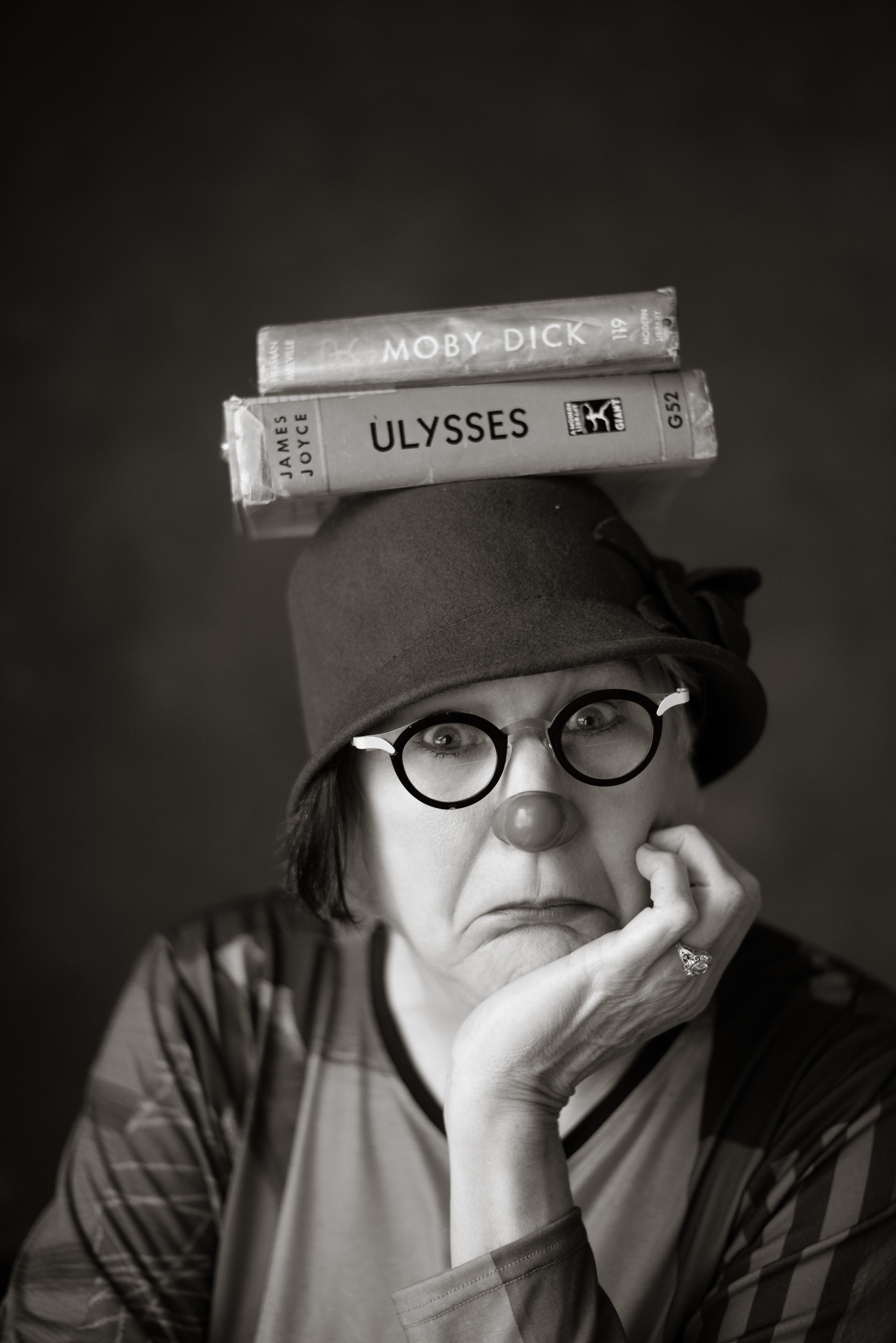 Black and white headshot of Victoria Millard with a clown nose on and books balancing on her head.